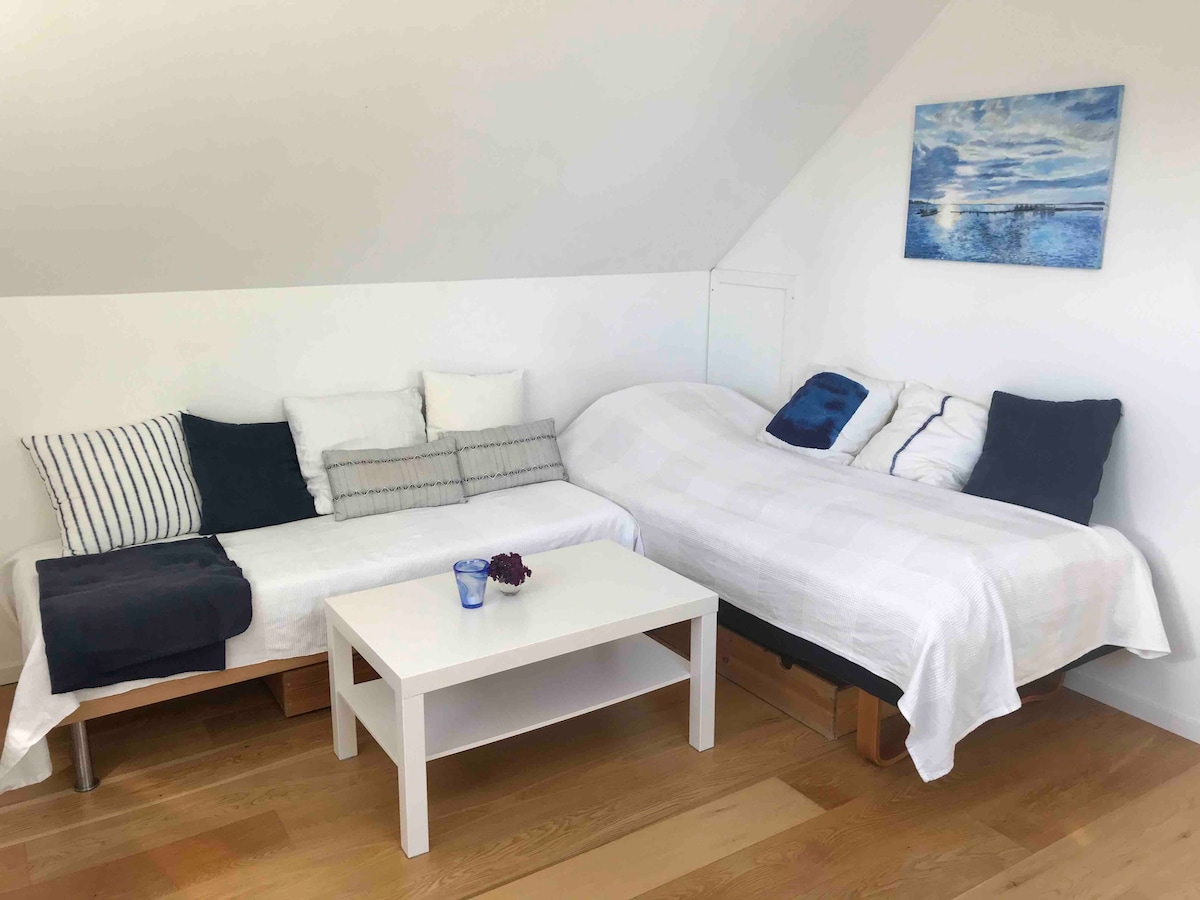 A well-lit sleeping area features two beds with white bedding, complemented by multiple pillows in varying shades. A small, modern white coffee table occupies the center of the space. A painting of a serene blue sky hangs on the wall, enhancing the room's calm ambiance.