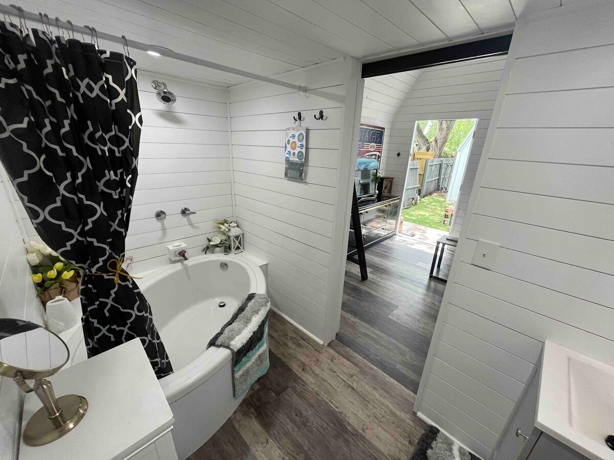 The bathroom features a corner soaking tub set against white shiplap walls. A dark patterned shower curtain adds a decorative touch. A small sink and mirror are positioned to one side, while natural light is visible from an adjacent room through a doorway.