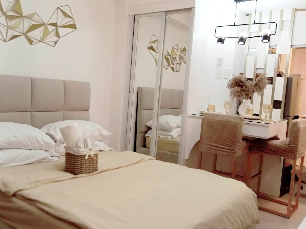 A modern bedroom features a spacious bed adorned with neutral-toned linens. A mirrored wardrobe reflects the room's design, while a chic table with chairs is situated nearby. Decorative geometric wall art adds a contemporary touch, enhancing the inviting space.