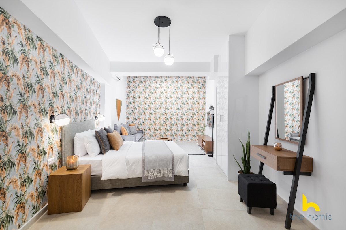 A spacious bedroom features a tropical-themed accent wall adorned with a floral print. A comfortable queen bed is flanked by two bedside lamps, with additional decorative pillows. A stylish mirror and a plant are positioned on a wooden console, complemented by neutral-toned bedding and flooring.