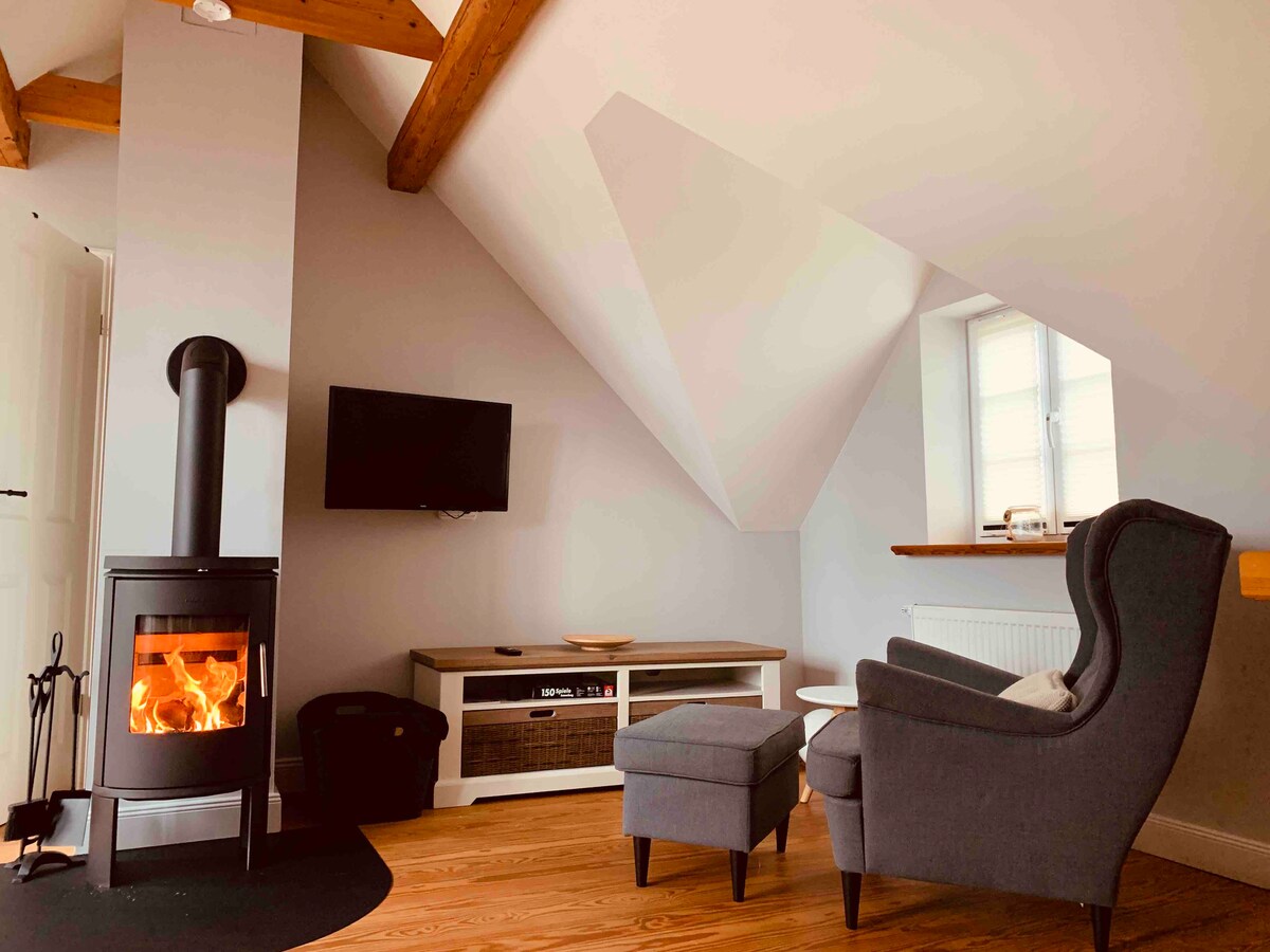 A cozy living area features a wood stove providing warmth. A grey armchair and a matching ottoman are situated next to a low wooden table. A wall-mounted television is placed opposite a white cabinet with storage. Natural light enters through a window set in a sloped ceiling.