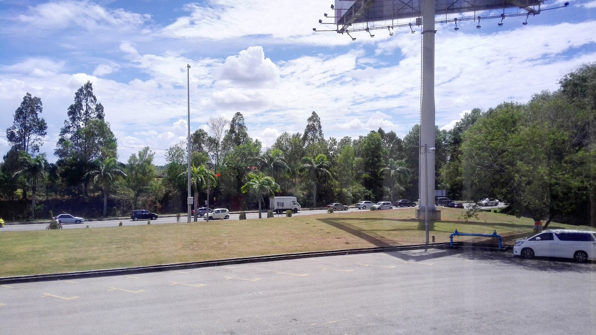 A view of the outdoor space reveals a grassy area bordered by trees and palm plants. A few parked vehicles are visible alongside the road, while a billboard stands in the foreground. The sky is bright with scattered clouds.