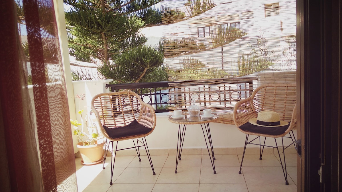 A cozy balcony features two rattan chairs and a small table, set against a backdrop of greenery. Sunlight filters through sheer curtains, creating a warm ambiance. Potted plants enhance the inviting space, perfect for enjoying morning coffee or evening relaxation.