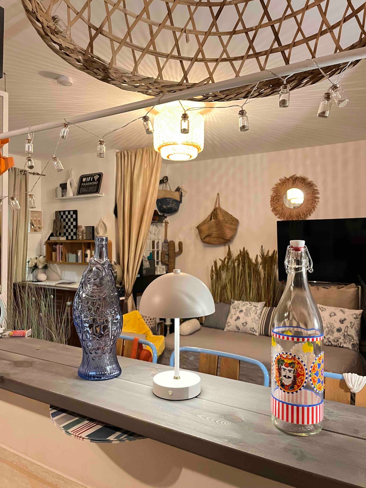 A stylish living area is presented, featuring a comfortable gray sofa and decorative wall elements. A colorful patterned glass bottle and a modern lamp are positioned on a countertop, illuminated by soft lighting from a pendant lamp above. Decorative plants add a natural touch to the space.