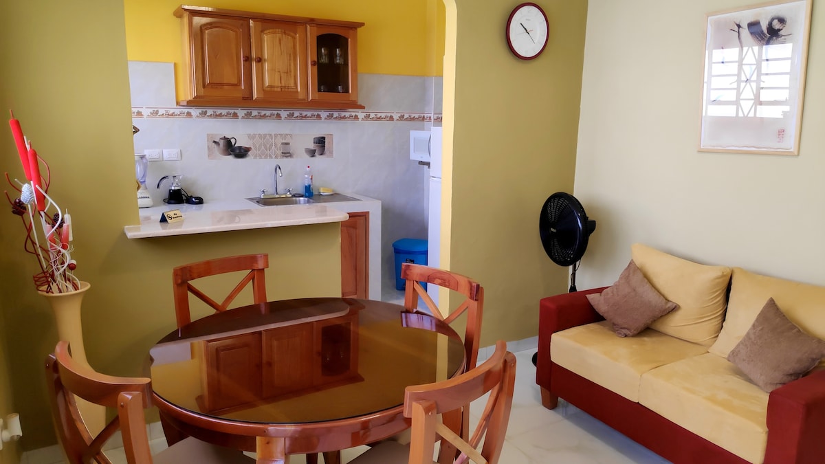 An inviting dining area features a round wooden table surrounded by four chairs. A cozy sofa with cushions is placed in the corner. A well-equipped kitchen is visible in the background, highlighted by wooden cabinetry and a clock on the wall.