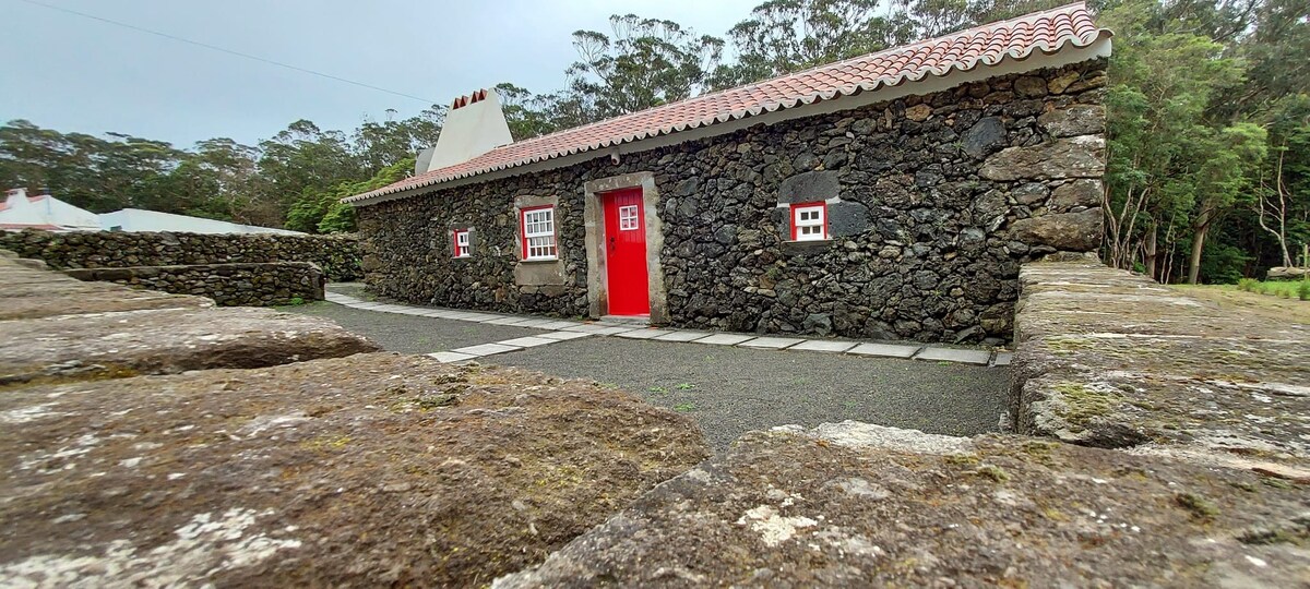 A traditional stone cottage is characterized by its rugged exterior and a vibrant red door. The structure is surrounded by a low stone wall, with a pathway leading to the entrance. Lush greenery is visible in the background, enhancing the natural setting.