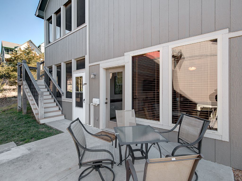 An outdoor patio area features a glass table surrounded by four metal chairs, providing a space for dining or relaxation. The entrance to the lower level of the cabin is visible, along with stairs leading up to the upper floor. Windows allow natural light to illuminate the interior.