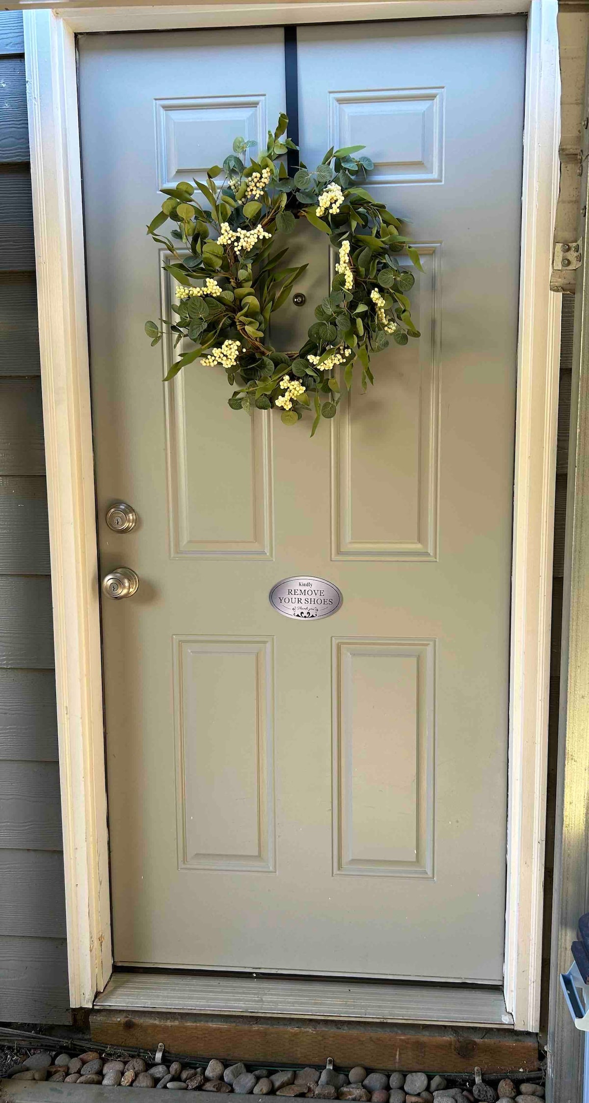 The entrance door features a soft green color and is adorned with a circular floral wreath. A small, circular plaque is mounted next to the door handle, providing welcoming information. The area is bordered by smooth pebbles, enhancing the outdoor ambiance.