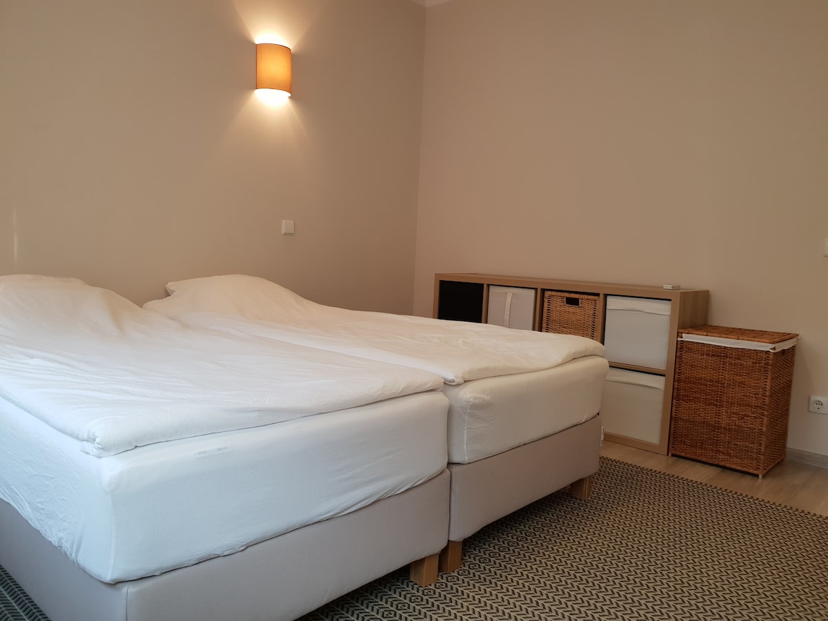A bright bedroom features two spacious beds that can be pushed together, with light walls and warm lighting from a wall fixture. A stylish woven basket and a dresser with closed storage are visible, contributing to the room's simplicity and functionality.