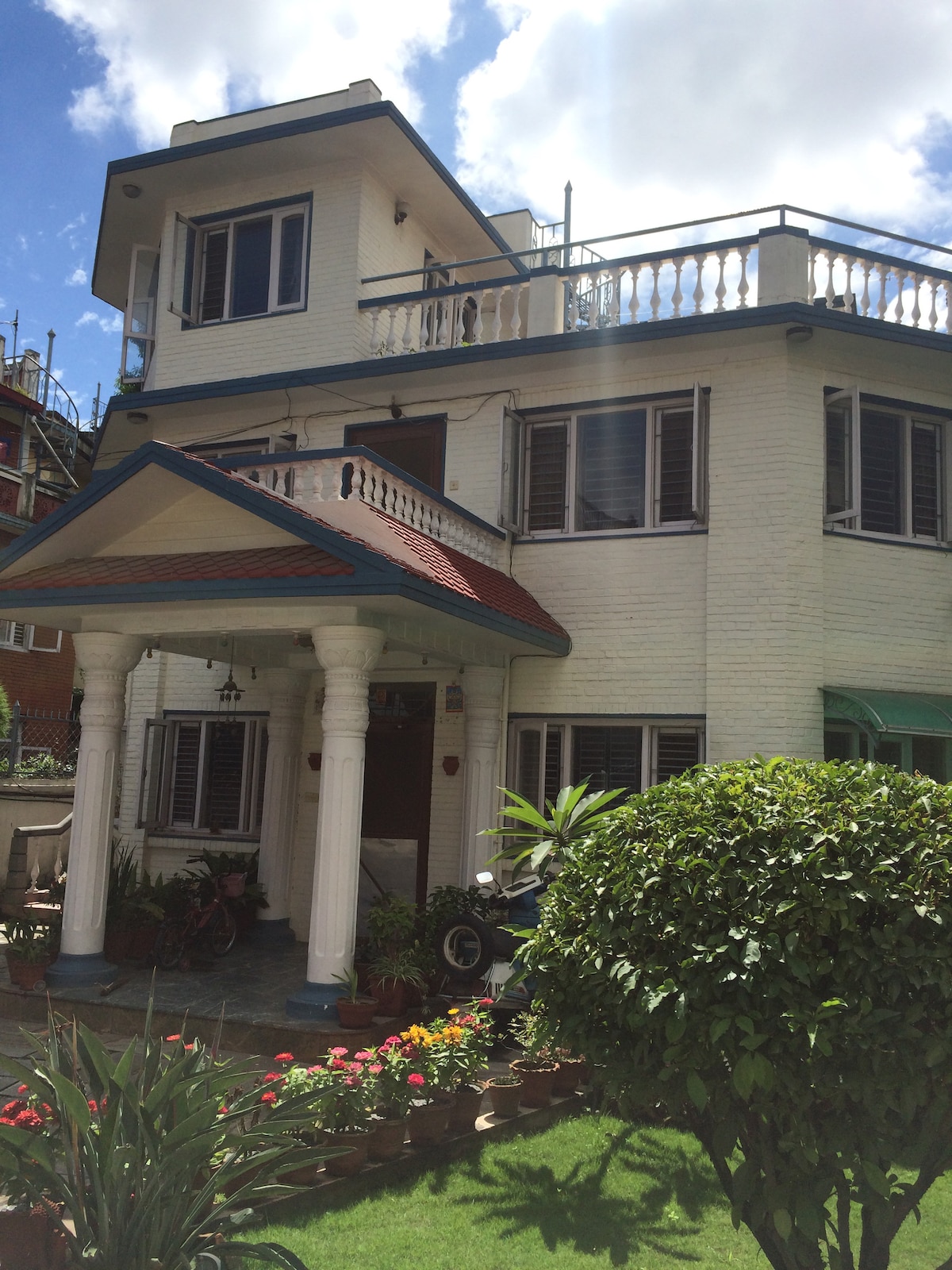 The exterior of a two-story house is seen, featuring a welcoming entrance with decorative columns and a tiled porch. Windows are visible on both levels, framed by greenery and flower pots in the garden, adding a touch of color to the front yard.