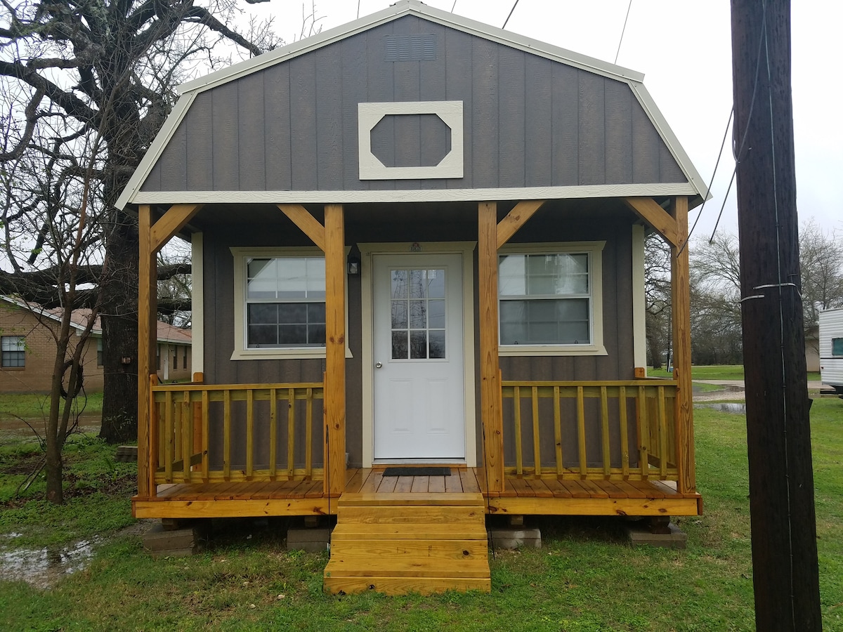 A small, rustic cabin features a wooden porch with a sturdy railing. The white front door is framed by two large windows, allowing natural light to enter. The exterior is clad in dark brown paneling, complemented by a light-colored trim.