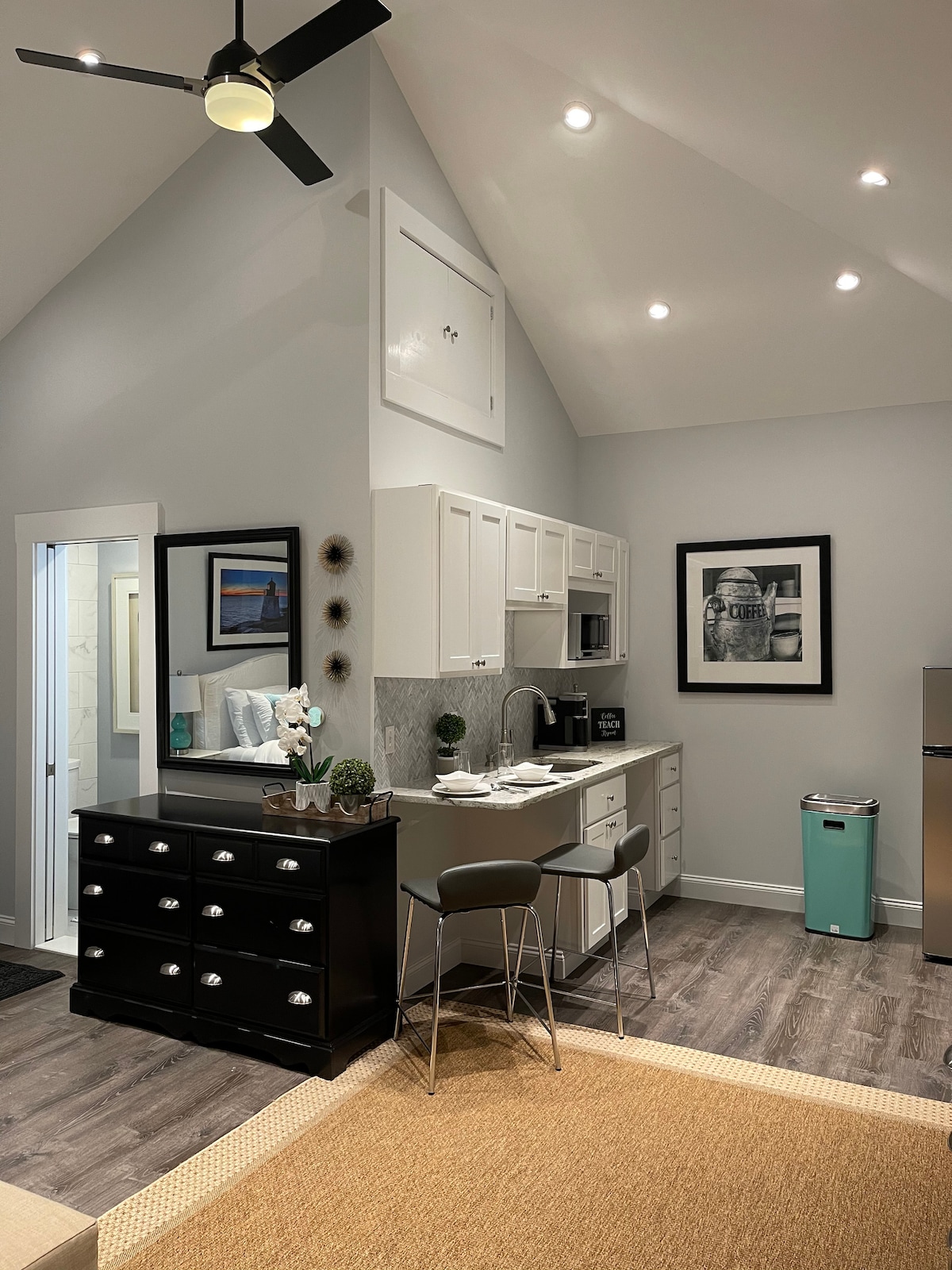 An open kitchen space features a high, sloped ceiling with recessed lighting. White cabinets and modern appliances are visible, along with a sleek black dresser and bar stools. A framed artwork adorns the wall, complementing the clean and organized layout.