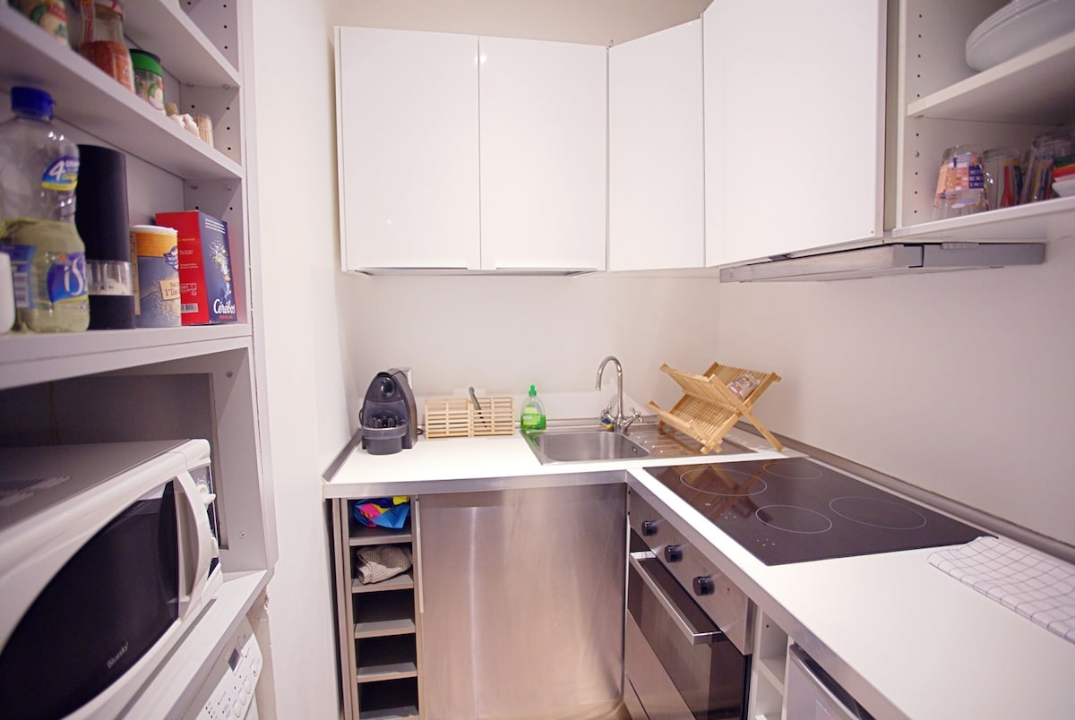 A modern kitchen is showcased with sleek white cabinetry and stainless steel appliances. An induction cooktop and sink are positioned on the countertop. Shelving holds kitchen utensils and supplies, while a microwave and refrigerator are visible in the space.