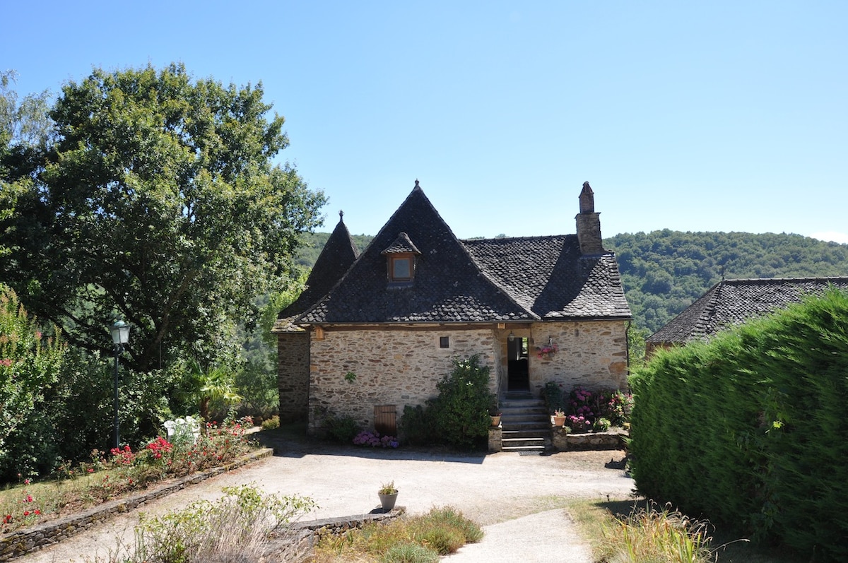A charming stone house features a distinctive steep roof and is surrounded by lush greenery. A gravel pathway leads to the entrance, framed by vibrant flowers and mature trees. The scenic hillside is visible in the background, enhancing the serene setting.