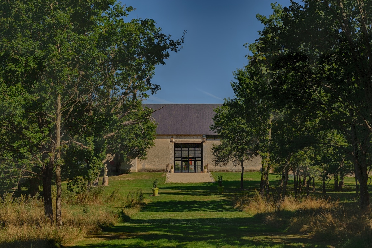 The image features a large, rustic building framed by tall trees and lush greenery. A spacious lawn leads to the entrance, highlighted by large glass doors that provide a welcoming view into the interior. The clear sky can be seen in the background.