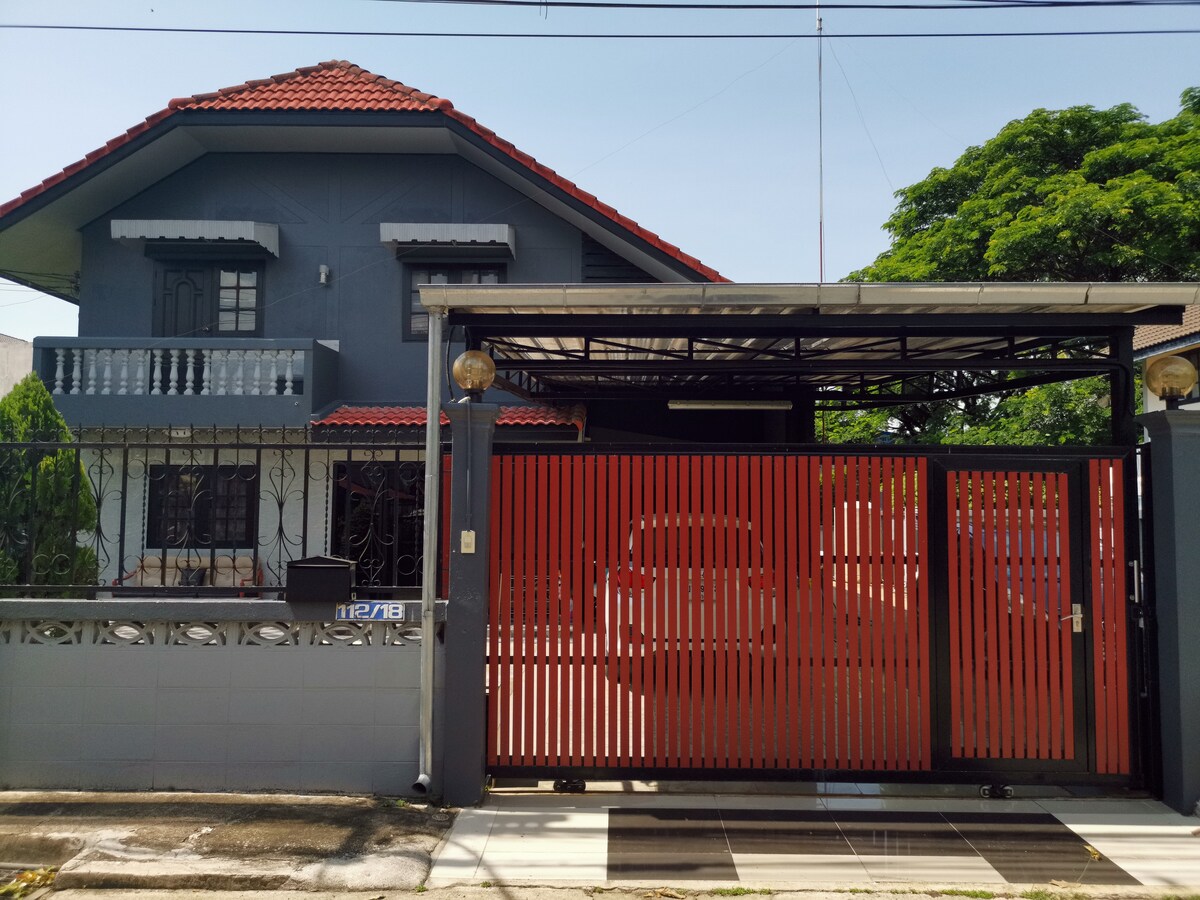 A single house featuring a grey exterior with red accents, highlighted by a distinctive orange gate. The two-story structure is surrounded by greenery, with a front porch visible. The residential layout offers a welcoming entrance, complementing the peaceful neighborhood setting.