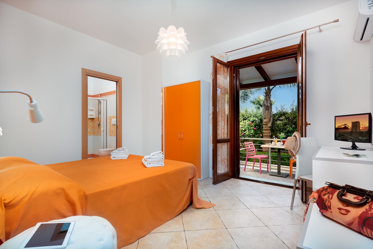 A modern bedroom features an orange bedspread draped over a spacious bed. Natural light enters through open wooden doors leading to an outdoor area with a table and chairs. A television and stylish lamp are visible on a desk, while a bathroom area is partially seen.