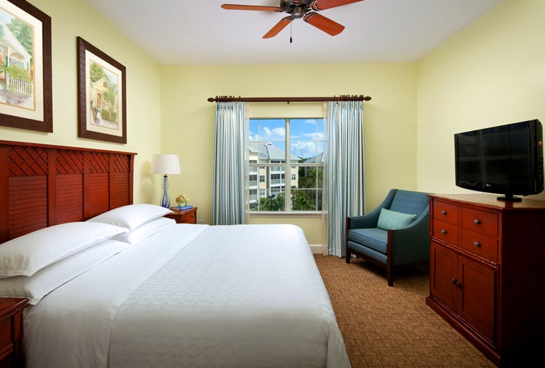 A bright and welcoming bedroom features a spacious bed with crisp white linens. A window allows natural light to fill the room, while a wooden dresser and a cozy chair add functional elements. Soft blue curtains frame the window, complementing the light yellow walls.