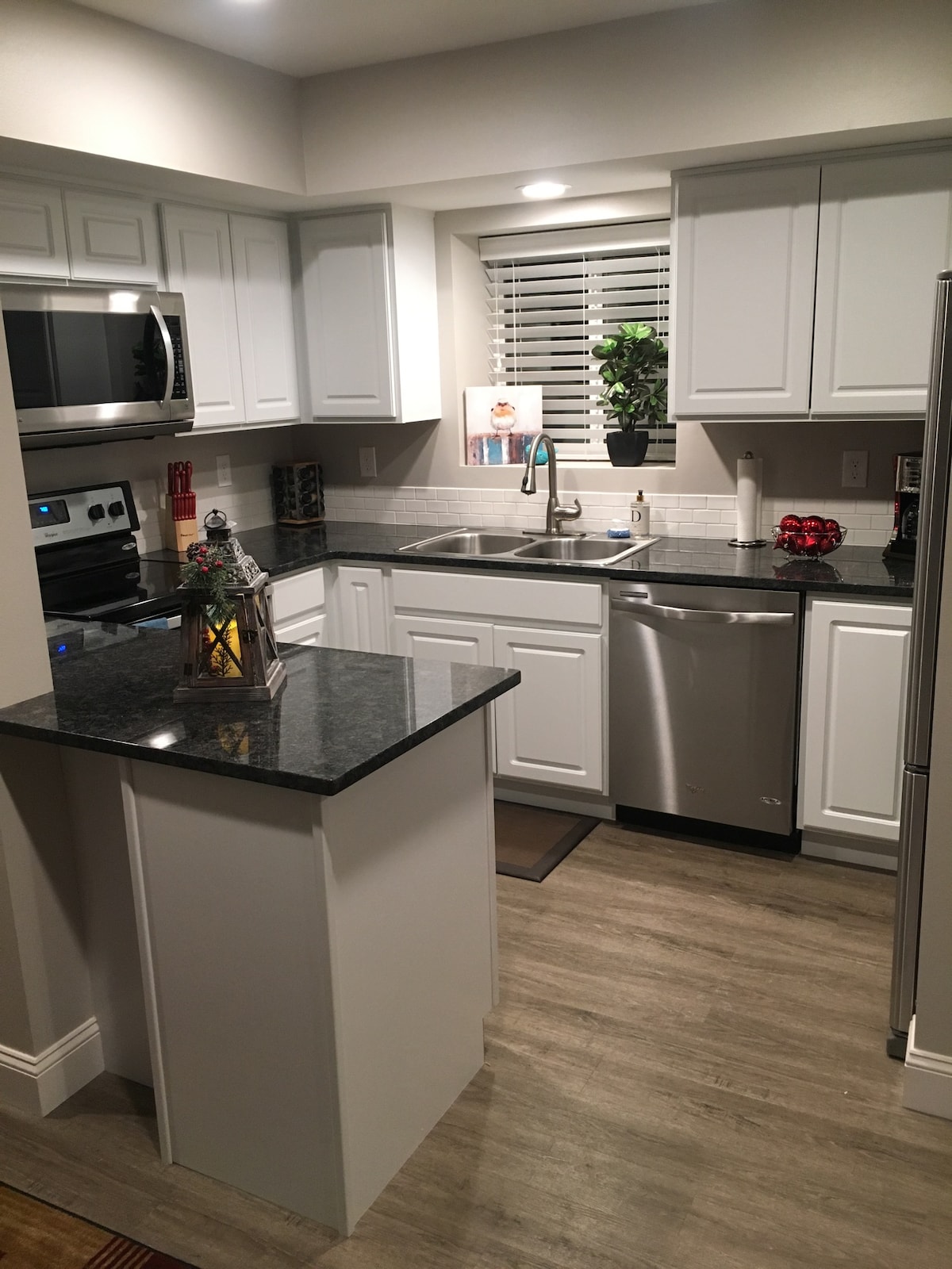 A modern kitchen features a stainless steel refrigerator and dishwasher, with a sleek gray countertop complemented by white cabinetry. A window allows natural light to illuminate the space, while a decorative plant and a lantern add a touch of homely warmth.