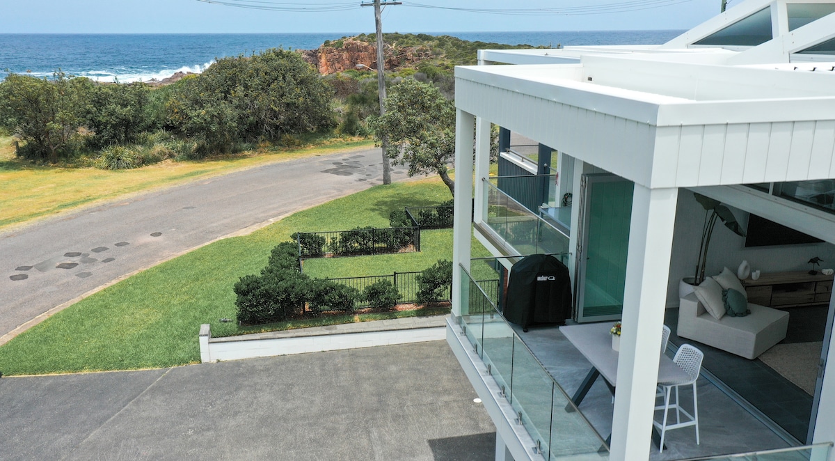 The property features a spacious balcony with a view of the ocean, equipped with a barbecue. The surrounding landscape includes well-maintained grass and low shrubs, visible from the modern structure. A quiet street leads to the beach, enhancing the coastal ambiance.