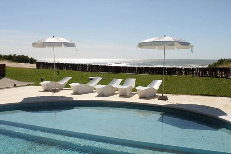 A sunlit pool area features white lounge chairs positioned near a tranquil blue pool. Two large umbrellas provide shade, while a grassy area extends toward a beach with gentle waves visible in the distance.