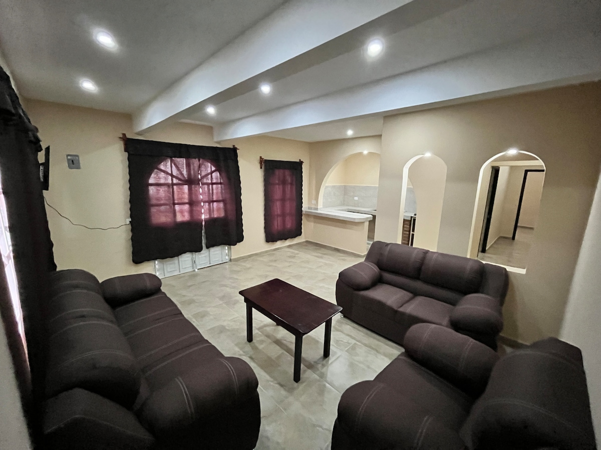 A spacious living area features two dark brown sofas arranged around a central coffee table. Natural light filters through large arched windows, adorned with dark curtains. The neutral wall tones add to the open feel of the room, which includes archways leading to adjacent spaces.
