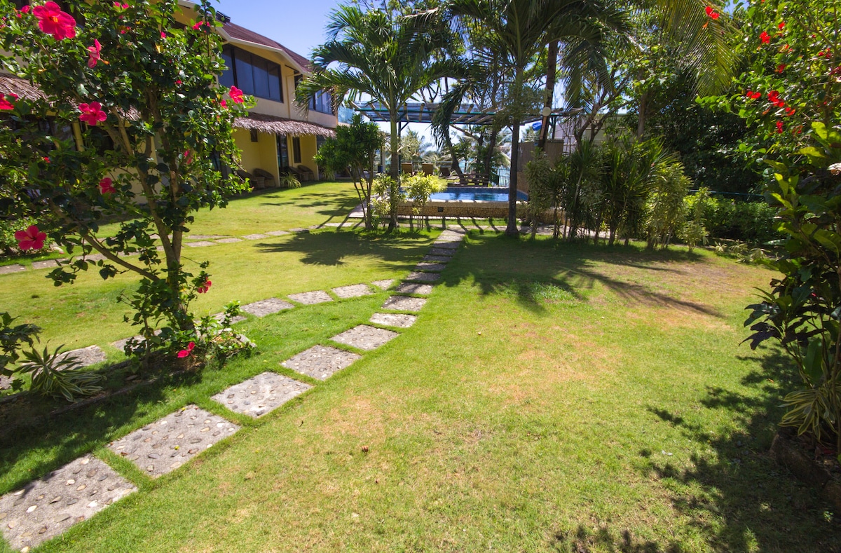 A lush garden is showcased, featuring a well-maintained lawn and vibrant flowers. Stone path pavers meander through the greenery, leading toward a shared dipping pool surrounded by tropical plants and palm trees. The bright blue sky provides a pleasant backdrop to the tranquil scene.