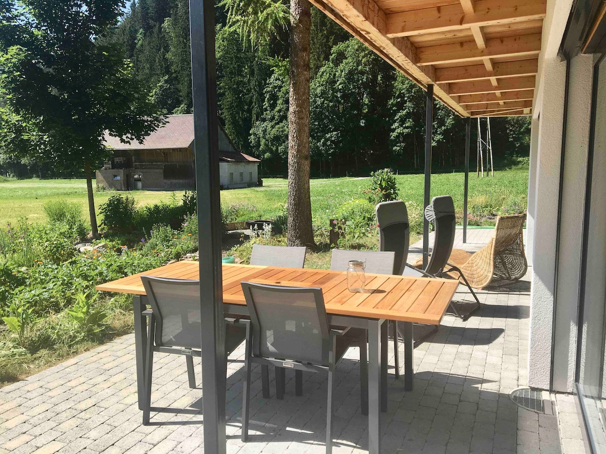 A spacious outdoor patio features a wooden dining table surrounded by comfortable seating. Lush greenery and trees create a serene backdrop, while a rustic building is visible in the distance, enhancing the natural ambiance.