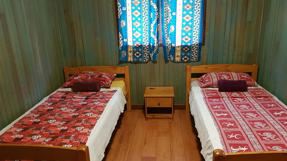 A simple guest room features two wooden beds, each adorned with colorful bedding in red and patterned textiles. A small wooden nightstand sits between the beds. Natural light filters through two bright blue curtains, illuminating the wood-paneled walls.