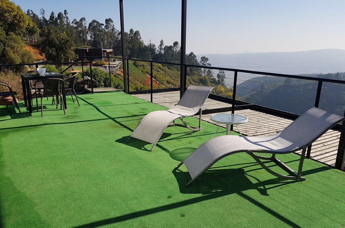 A spacious terrace is presented, featuring two lounge chairs and a glass table, all set on green artificial grass. The background reveals a scenic view of rolling hills and trees under a clear sky, emphasizing the serene outdoor space.