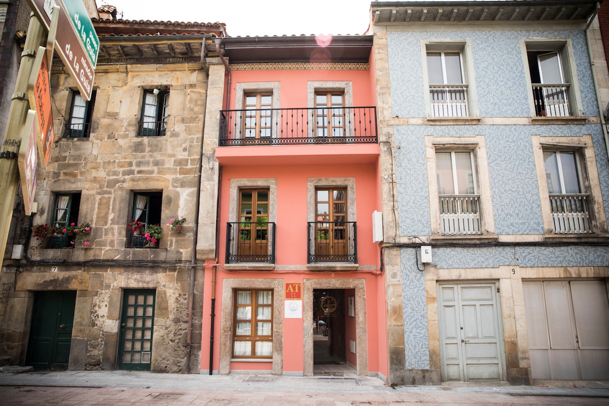 A charming three-story building is presented in the image, featuring a pink facade with decorative balconies. The adjacent structures include stone and patterned blue textures, creating a blend of architectural styles. Flowers adorn the windows, contributing to the overall aesthetic of the street.