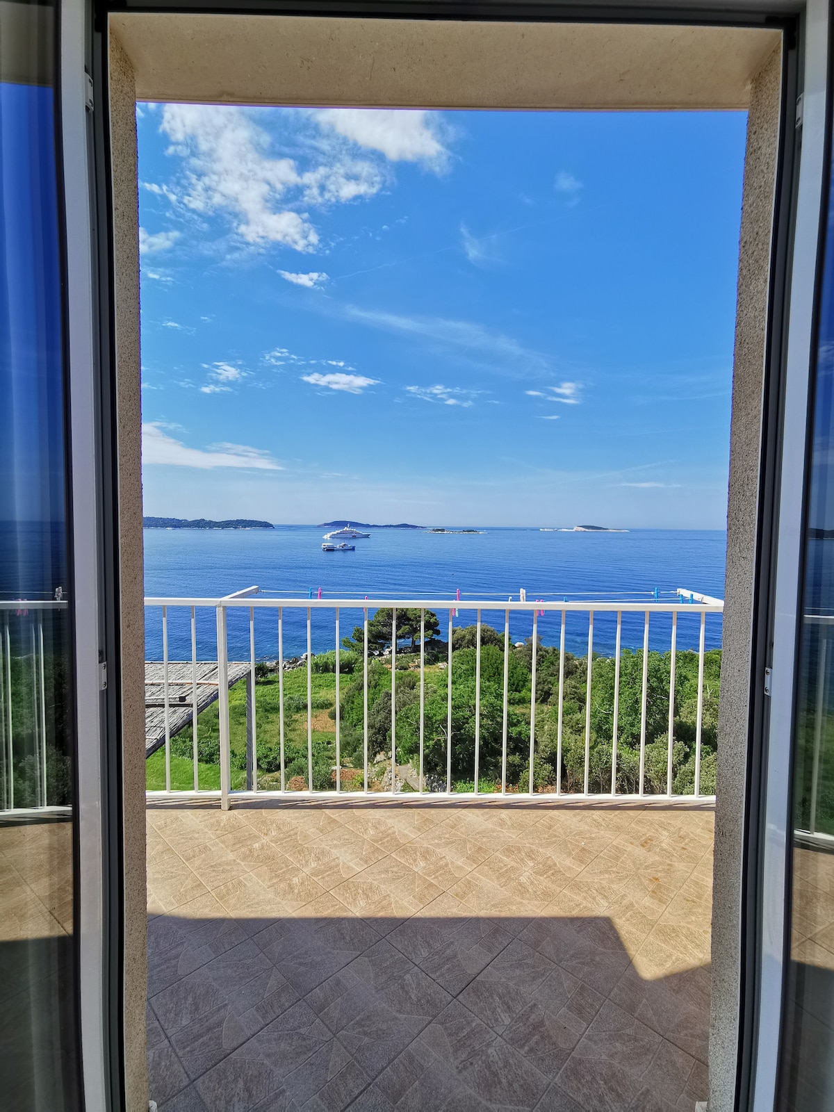 A balcony view is captured, showcasing the expansive blue sea and sky. The scene includes distant islands and a boat traversing the water. The glass doors frame the view, inviting natural light into the interior space.