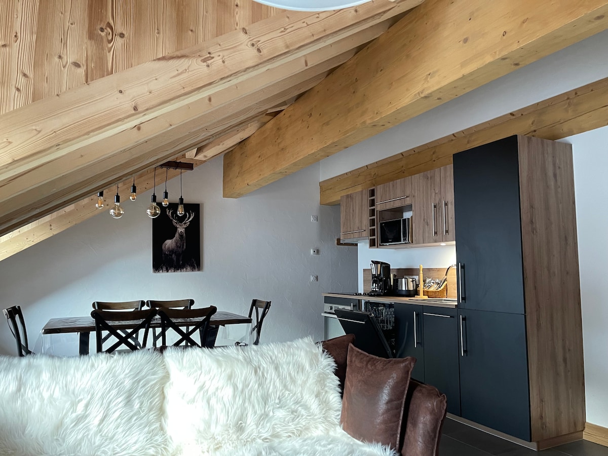An open living space features wooden beams and a modern kitchen area. A cozy sofa with a white throw faces a wooden dining table. Pendant lighting hangs above the dining area, while a wall display includes an artwork of a deer, enhancing the alpine décor.