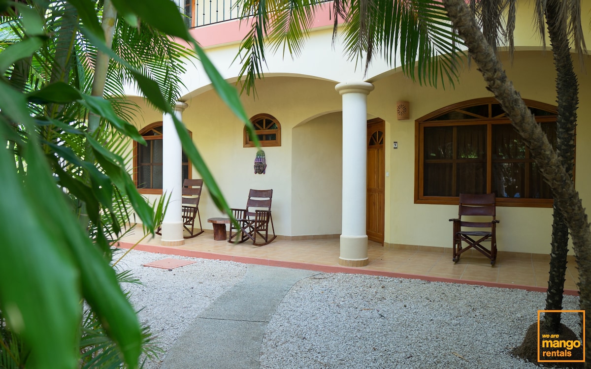 A charming entrance area features a pathway lined with white pebbles and tropical foliage. Two wooden rocking chairs are positioned on a covered porch, inviting relaxation. Large arched windows provide ample natural light to the space, enhancing the rustic appeal.