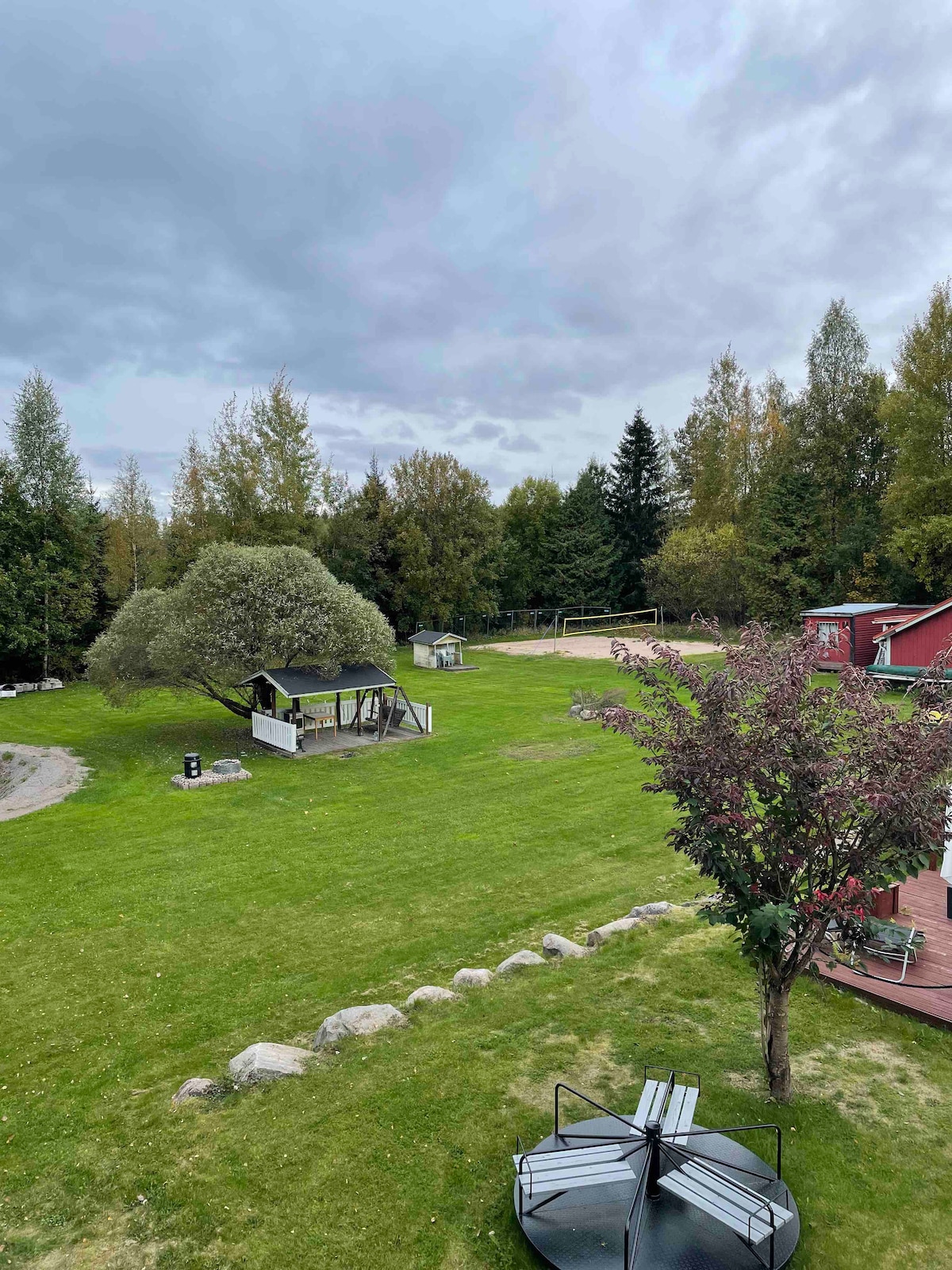 A large green lawn is bordered by trees and boulders. A covered grill area is visible alongside a playground. A small structure can be seen in the background, with an open space suggesting recreational use. The sky appears overcast, adding depth to the landscape.