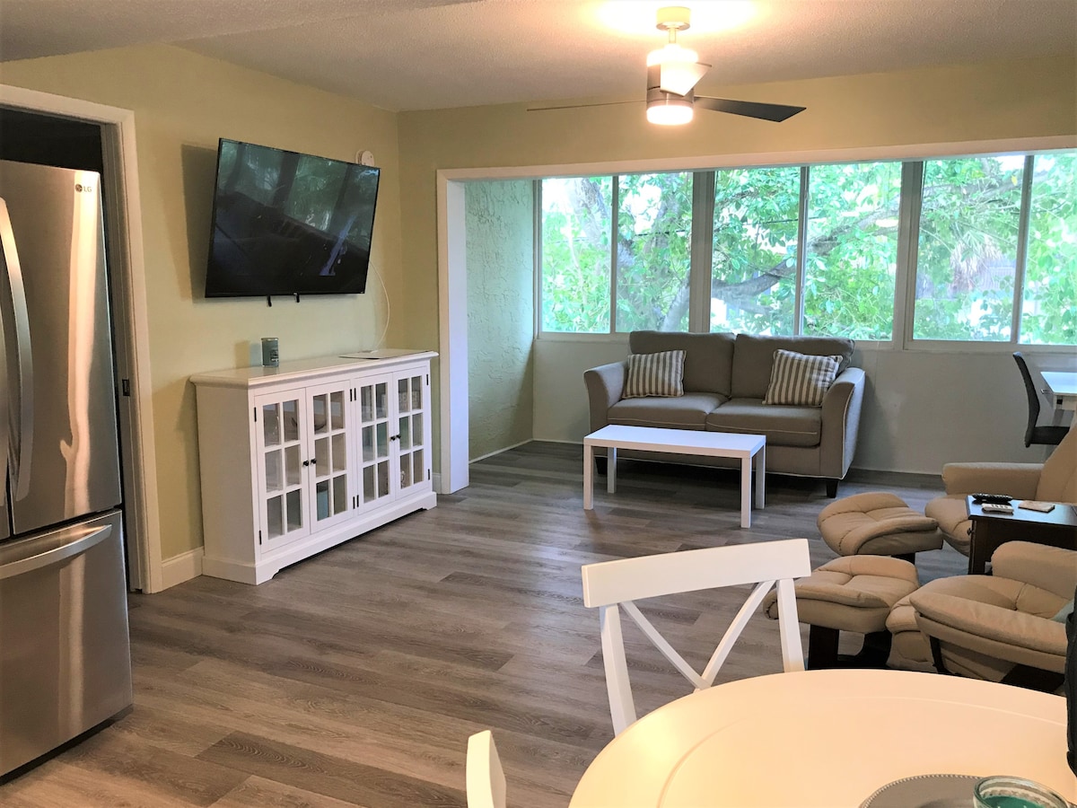 A bright living area is presented, featuring a comfortable sofa with decorative cushions, a coffee table, and a large TV mounted on the wall. Windows invite natural light, creating an airy atmosphere. A dining table and modern seating are visible in the background.