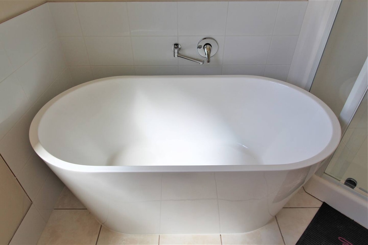 A free-standing bathtub is positioned in a bright bathroom, featuring smooth curves and a glossy white finish. The surrounding tiles create a neutral and clean backdrop. A shower area is visible to the side, with modern fixtures providing a contemporary touch.