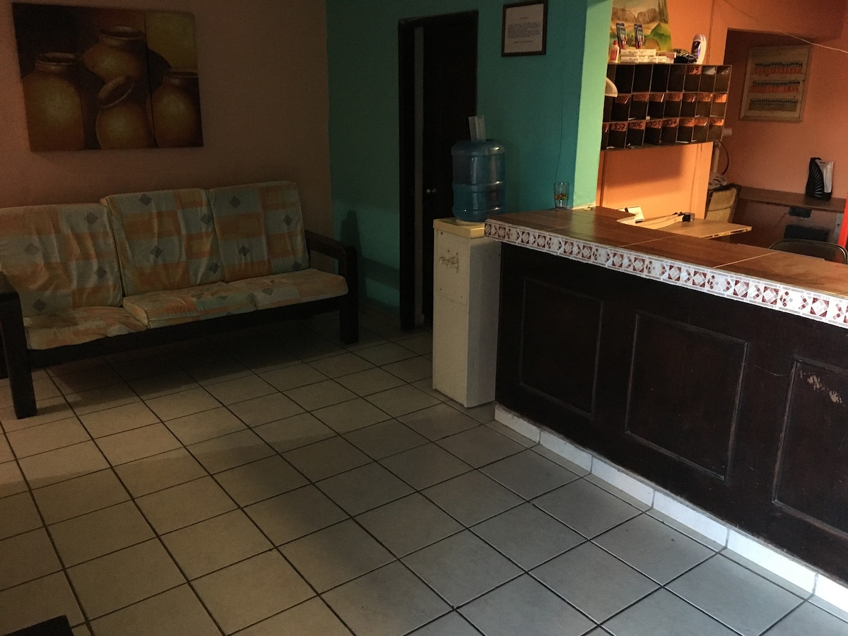 A reception area is featured, showcasing a simple counter with a decorative edge. A water dispenser stands nearby. A couch with a patterned cover is positioned against a wall adorned with artwork. The tiled floor contributes to a clean, functional environment.