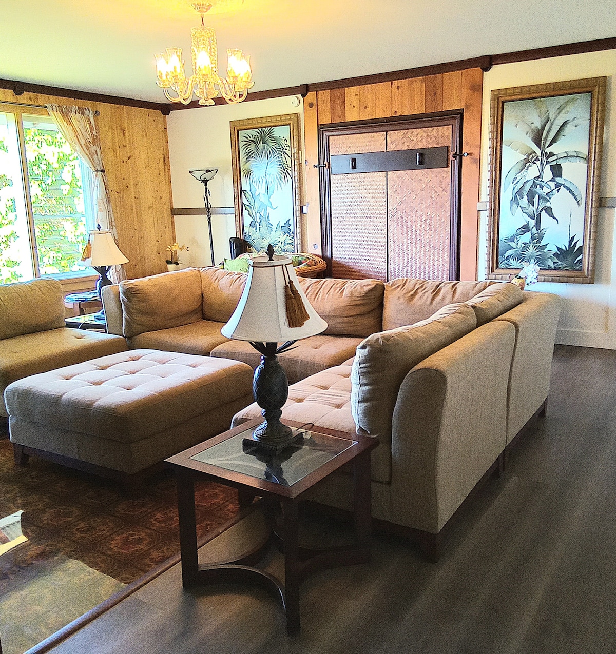 A spacious living area features a large L-shaped sofa with neutral upholstery, complemented by a glass-topped side table. Decorative artwork with tropical themes adorns the walls, while a chandelier provides soft illumination. Large windows allow natural light to filter in, enhancing the open ambiance.