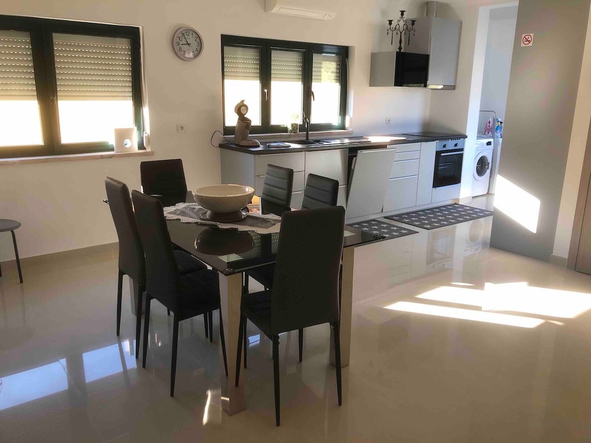 A bright and modern kitchen and dining area is featured, showcasing a sleek table surrounded by six chairs. Large windows provide ample natural light, reflecting off the glossy tiled floor. The kitchen includes contemporary appliances, while a washing machine is visible in a corner.