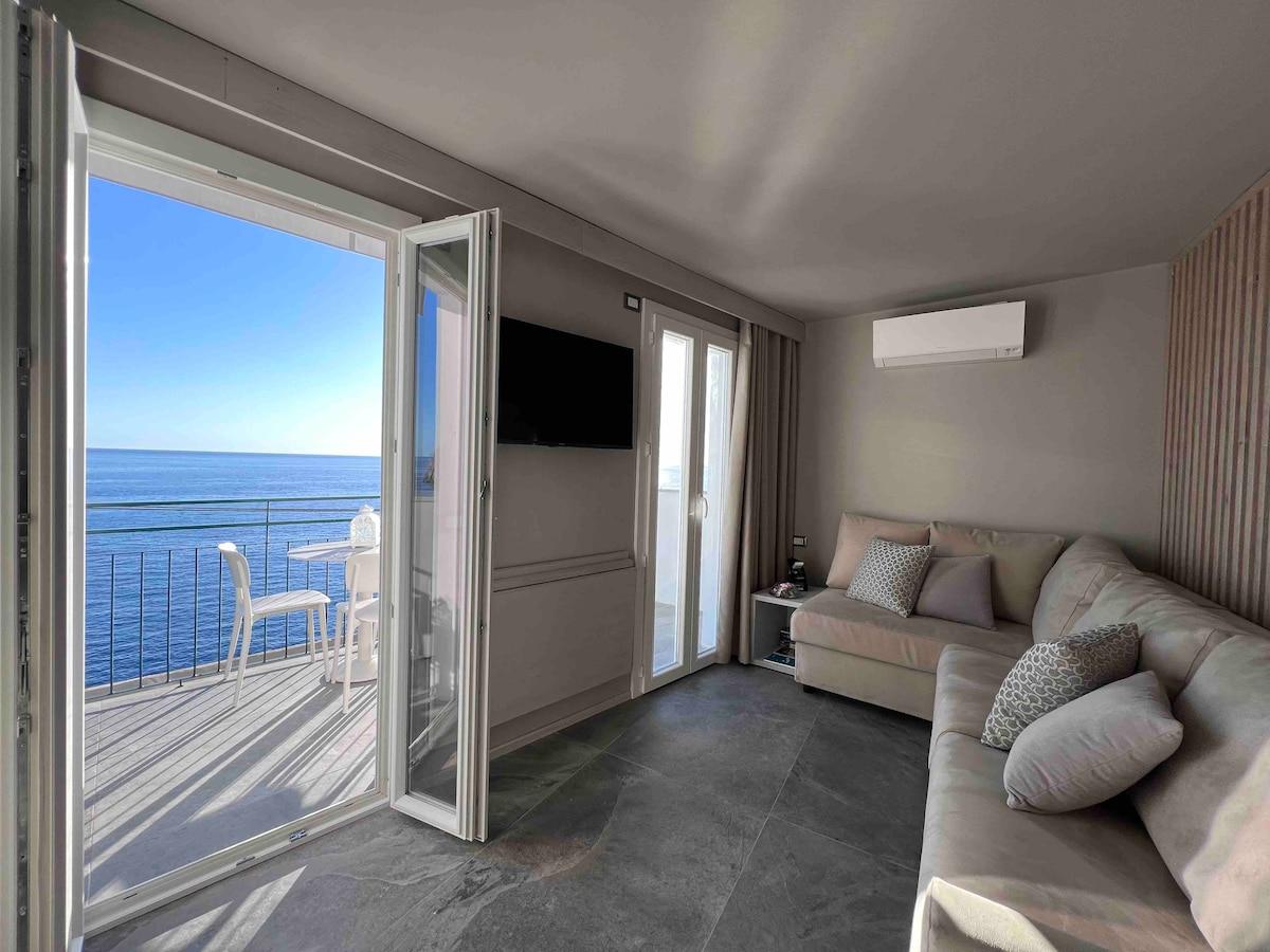 A bright living area features a comfortable sectional sofa adorned with soft pillows. Large glass doors open to a balcony with a view of the sea, allowing natural light to illuminate the space. A wall-mounted television and air conditioning unit are also visible.