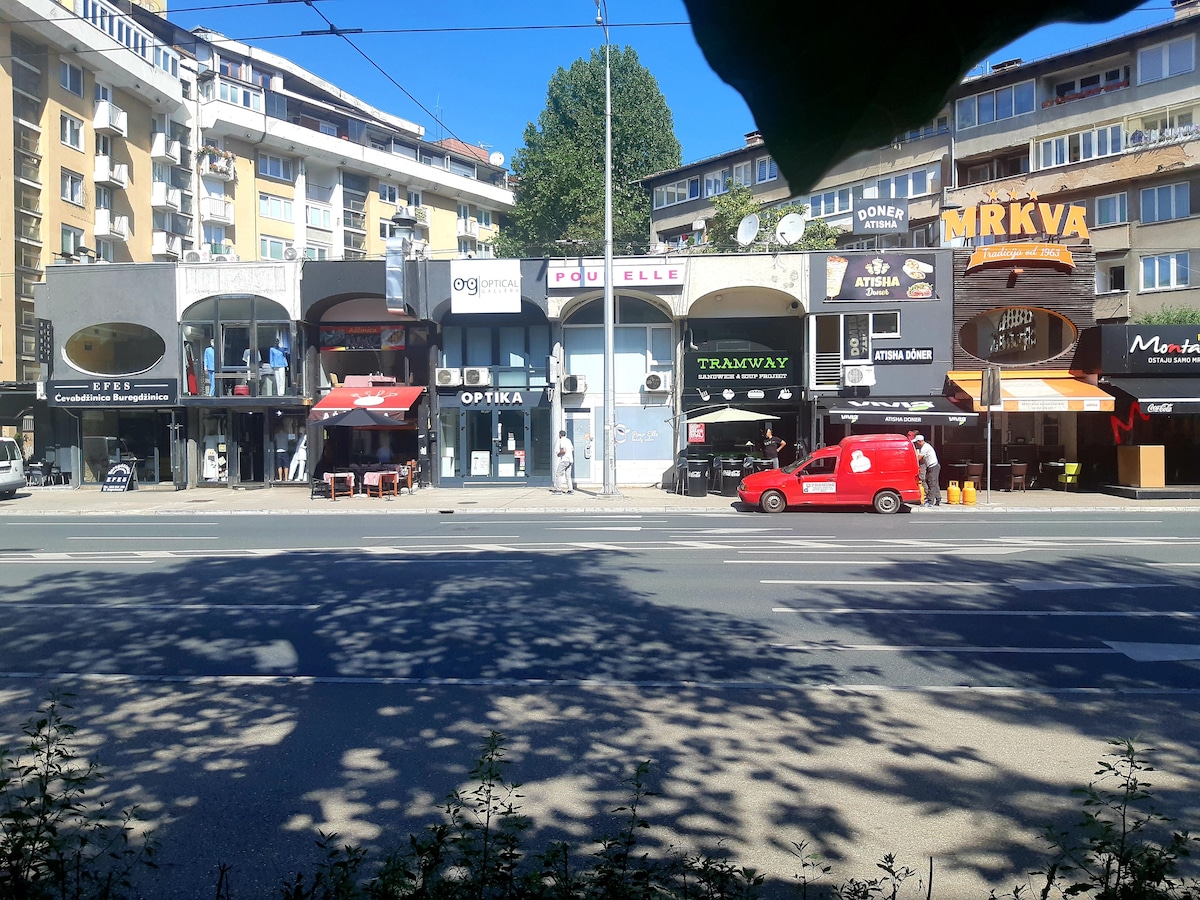 A bustling street scene captures a row of shops and cafes on a sunny day. The colorful awnings provide shade for outdoor seating, while a red delivery vehicle is parked along the curb. Visible trees line the street, and pedestrians can be seen enjoying the vibrant atmosphere.