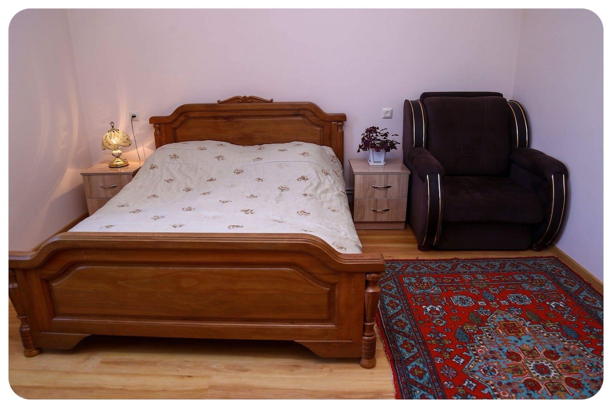 A wooden bed with a decorative headboard is positioned against a softly painted wall. Next to the bed, a bedside table is placed, accompanied by a stylish lamp. A plush armchair is situated in the corner, and a patterned area rug adds warmth to the wooden floor.
