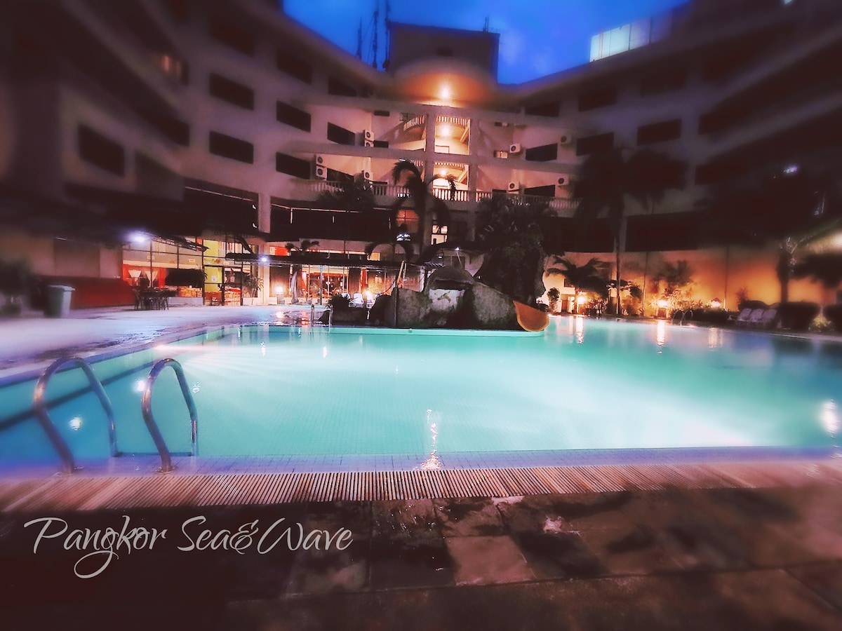 An outdoor swimming pool is illuminated with soft lighting, surrounded by tropical landscaping. The pool area features a gentle slope into the water and a sun deck lined with lounge chairs, creating an inviting space for relaxation. Nearby structures showcase balconies and windows, highlighting the surrounding atmosphere.