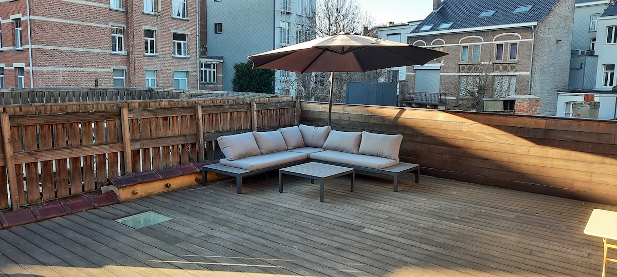 A spacious rooftop terrace is equipped with a comfortable seating area featuring a large sofa and a coffee table. An umbrella provides shade, while wooden decking surrounds the space. Views of neighboring buildings can be seen beyond the terrace's wooden perimeter.