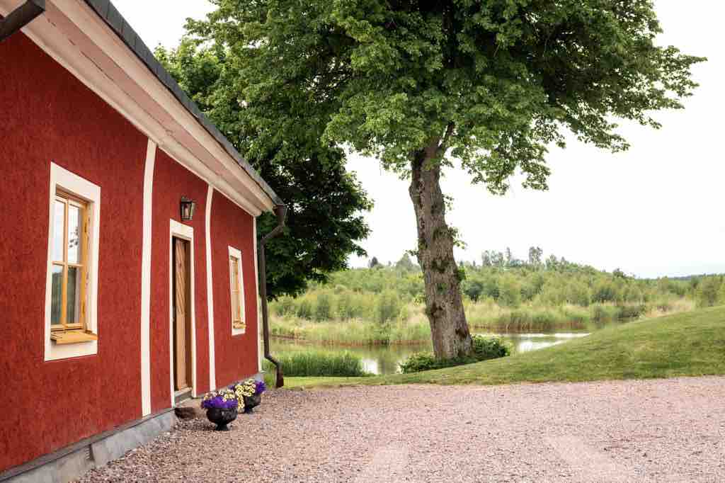 A charming red exterior of the building is highlighted by large windows and a welcoming door. The lush greenery and tranquil water are visible in the background, while flower pots line the walkway. The scene captures a peaceful outdoor ambiance.