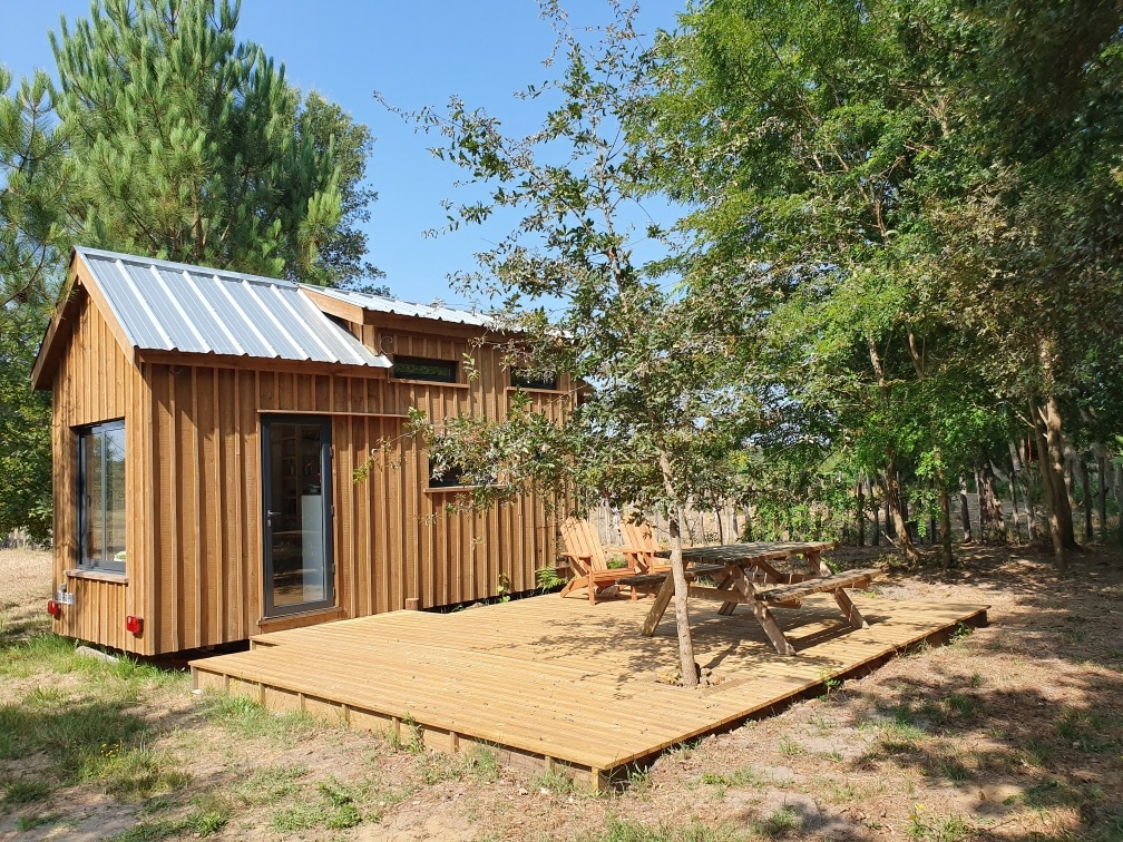 A wooden tiny house is surrounded by greenery, featuring large windows that allow natural light to fill the space. A spacious wooden deck includes seating areas and a picnic table, providing an ideal outdoor setting for relaxation.
