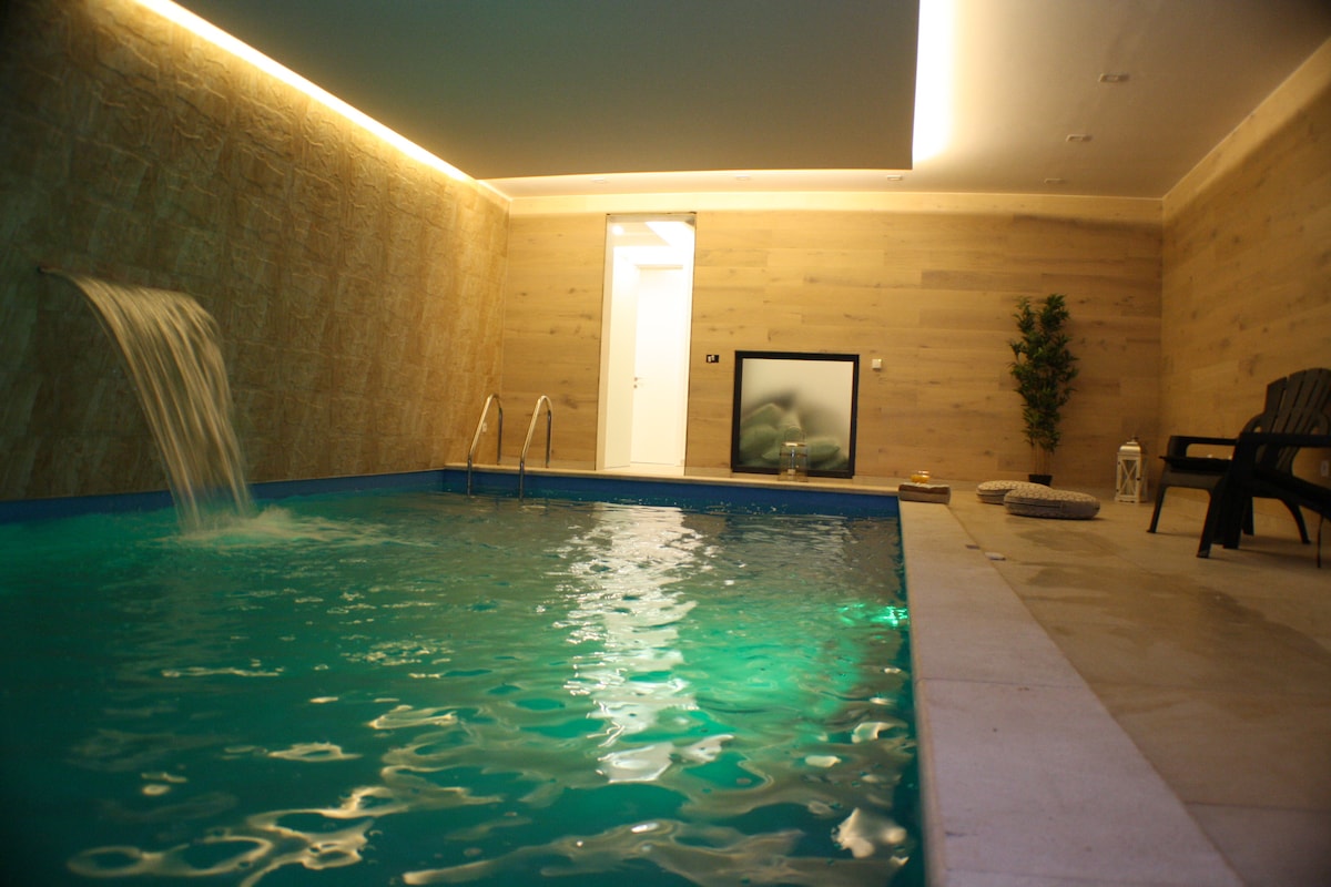 An indoor heated pool is depicted, featuring a soothing waterfall and illuminated turquoise water. The surrounding area includes light-colored wooden walls and modern seating. A large screen is visible on one side, and a potted plant adds a natural element to the tranquil setting.