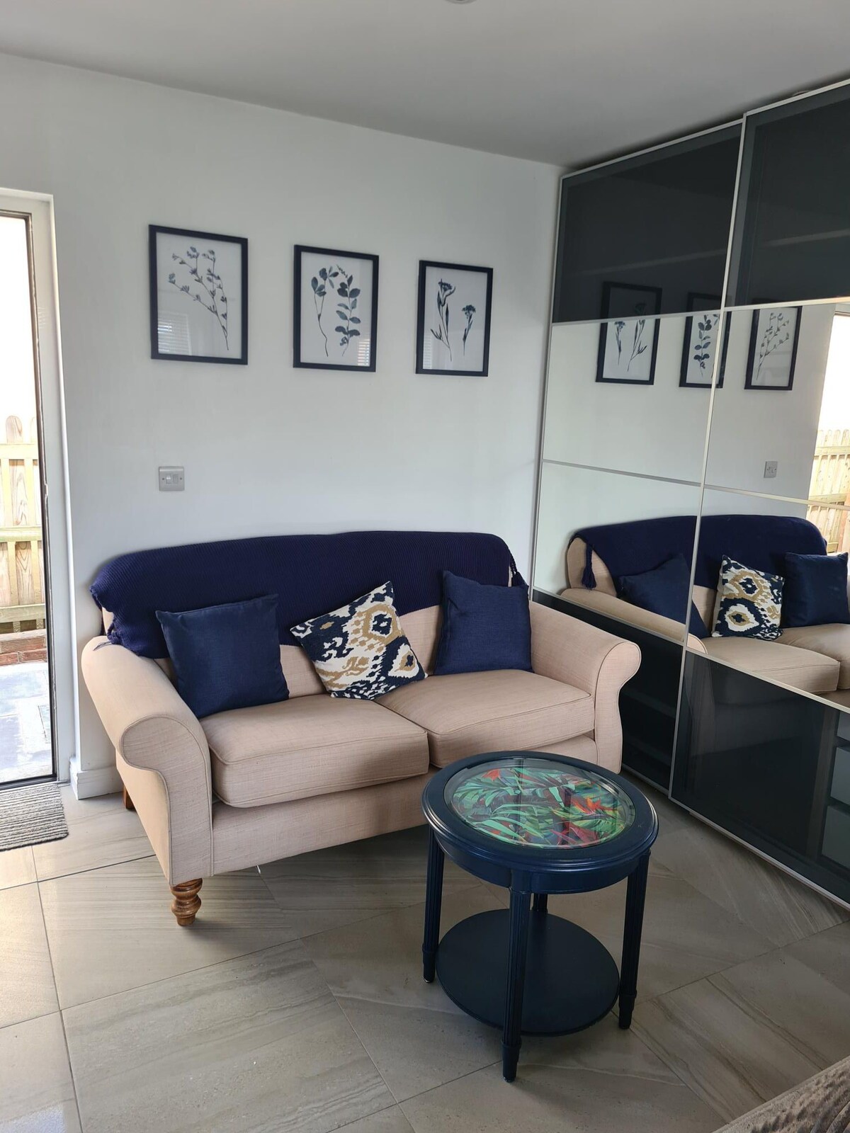 A cozy seating area is presented with a light-colored sofa adorned with navy and patterned cushions. A small, circular table rests beside the sofa, reflecting colorful artwork. The room is illuminated by natural light from an adjoining door, with a mirror enhancing the sense of space.