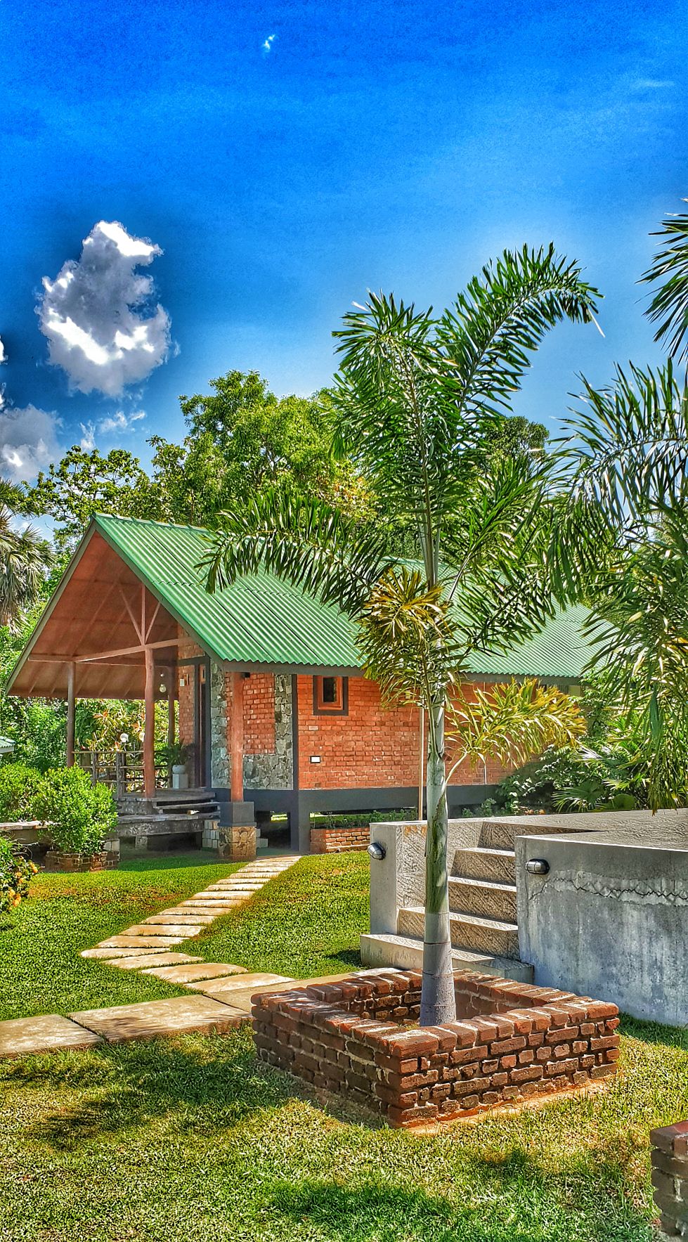 A charming villa is nestled among lush greenery, featuring a sloped green roof and brick exterior. A pathway of stepping stones leads to the entrance, framed by vibrant tropical plants. The clear blue sky adds a bright backdrop to this outdoor setting.